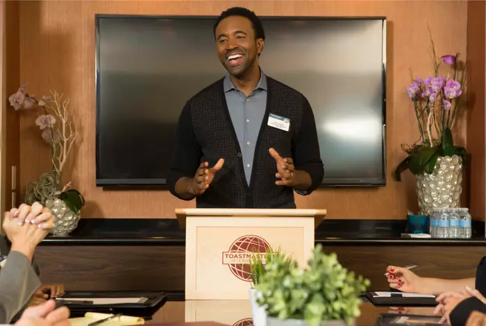 A Toastmasters member speaking from a lectern, smiling, while club members listen from the table around him.
